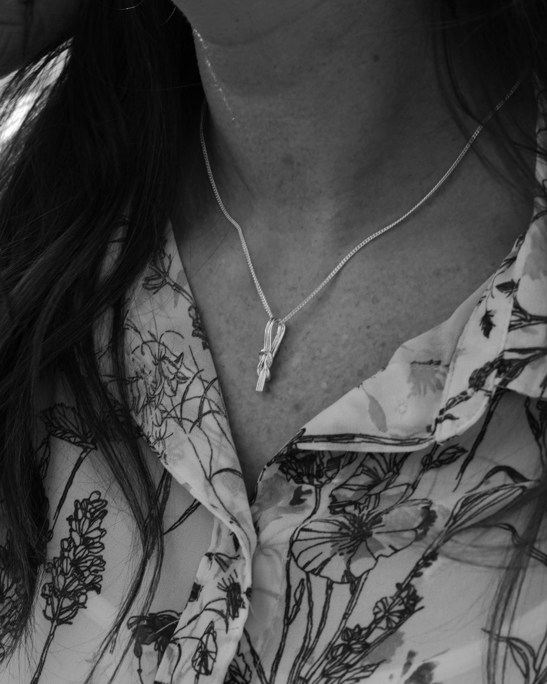 RÅBANDSKNOP halsband i silver med en knuten råbandsknop mot en blommig skjorta.