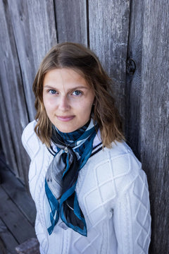 Model wearing the HEMMA HAMN scarf in blue tones against a weathered wooden fence